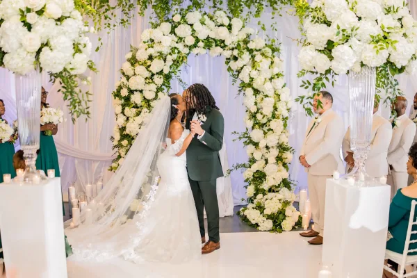 Ceremony kiss moment as the bride and groom kiss beneath a white floral arch, framed by white drapery in a bright indoor venue.