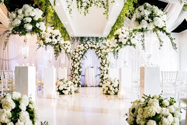 Ceremony setup with a floral ceremony arch and pillar candles lining the aisle, set against white draped walls and a glossy, bright indoor space.