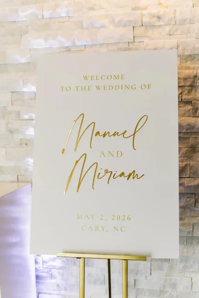 Wedding welcome sign with gold lettering, displayed against a stone wall, greeting arriving guests at the venue entrance.
