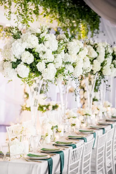 Tall wedding centerpieces elevate an elegant reception tablescape with white hydrangeas, beneath ceiling greenery and white drapery indoors.