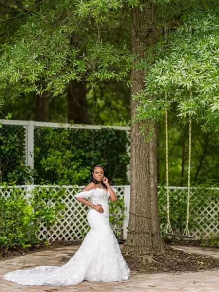 Bridal portrait of a bride under a tree, wearing an off-the-shoulder gown, standing on a stone patio beside a garden swing.
