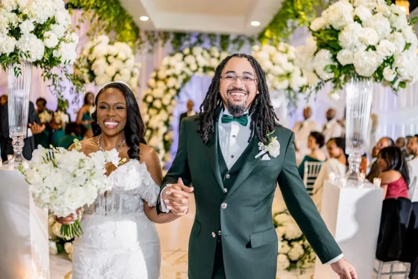 Ceremony recessional as a couple walking down the aisle, hands clasped and beaming, the groom in a green tuxedo along a draped indoor aisle.