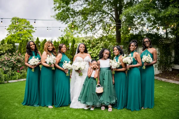 Bridal party portrait of a bride with bridesmaids and flower girls holding white bouquets in a garden with hedges and string lights.