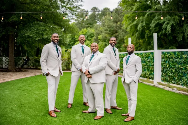 Groomsmen portrait of the groom with groomsmen in teal ties, relaxed on a garden lawn beneath string lights by a white fence.