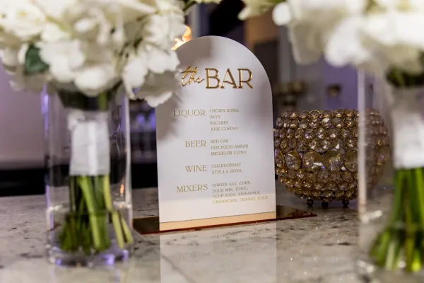 Wedding bar sign, a bar menu sign in white and gold, rests on a granite countertop beside a gold votive candle.