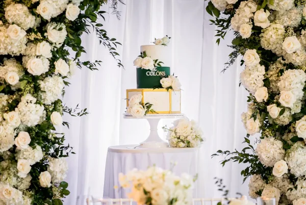 Wedding cake, a three-tier green and white cake adorned with white roses, framed by a soft white drapery backdrop