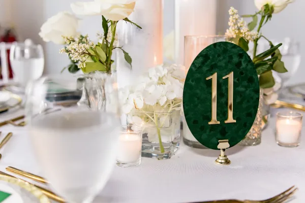 Wedding table number in green and gold on an oval sign, glowing beside candles for a minimalist, elegant reception table.