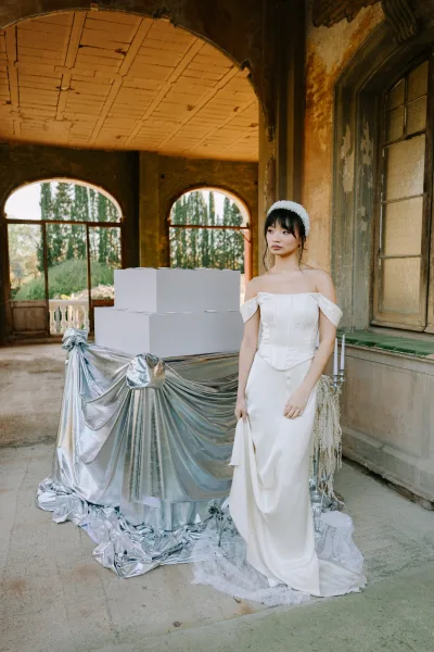 Bridal portrait of a bride in a strapless wedding dress with pearl headband, posed by stacked white boxes and taper candles outside a weathered building