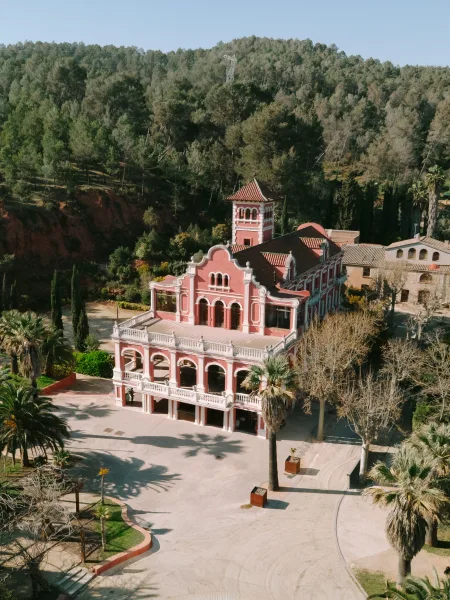 Wedding venue exterior with arched balconies and red roof tiles, a tower rising above a palm-lined courtyard against a forested hillside