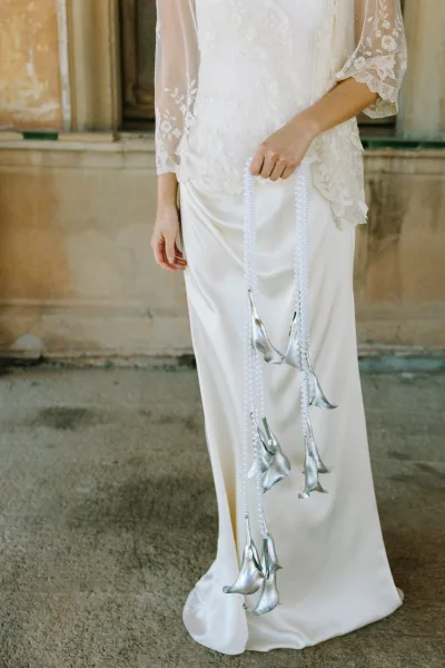 Wedding dress details featuring a lace bridal top with long sleeves, satin skirt, pearl strand, and silver calla lily bouquet by a stone wall
