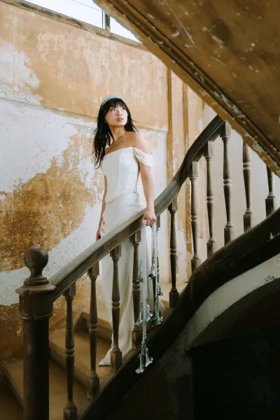 Bridal portrait of a bride on staircase in an off the shoulder corset wedding dress, wearing a tiara, lit by window light in an old building