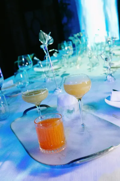 Reception tablescape with wedding cocktail table setting featuring coupe glasses, white plates, silver flatware, in a dim blue-lit room
