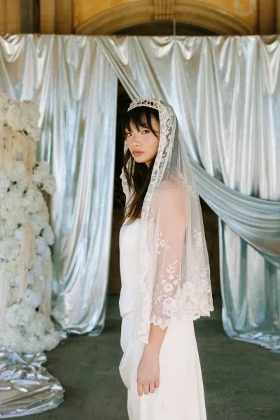 Bridal portrait of a bride wearing tiara with a lace veil, looking over her shoulder in a strapless dress by draped curtain backdrop