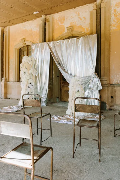 Ceremony backdrop with silver drape wedding backdrop, white floral pillars, and metal folding chairs before a weathered arched doorway porch
