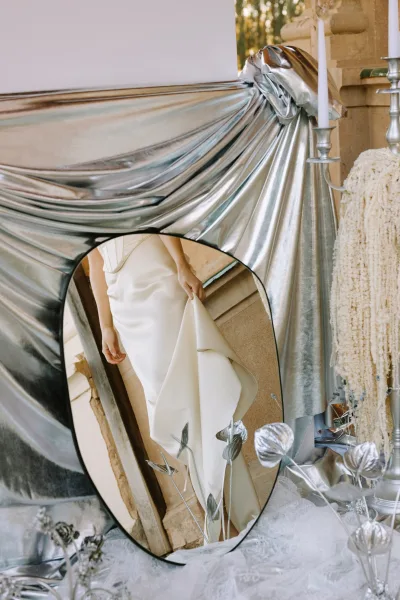 Wedding dress detail with satin wedding dress draped beside a mirror, silver candlesticks and taper candles against a stone wall backdrop