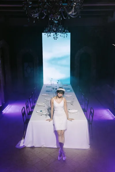 Bridal portrait of a bride in a short wedding dress and headband leaning on a banquet table with chandelier and foggy, dark reception room backdrop