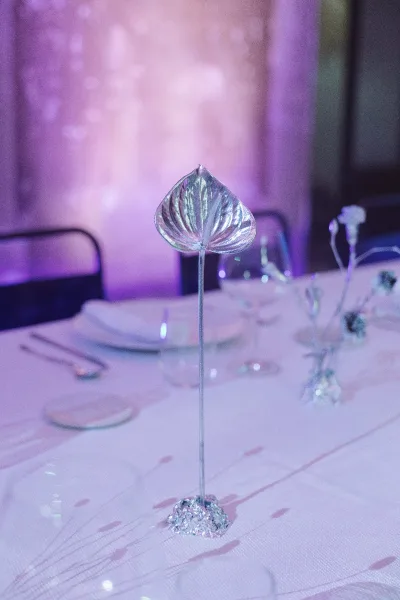 Wedding table centerpiece with silver leaf decoration on a clear stem and crystal base, set on pink linen with white plates and purple uplighting