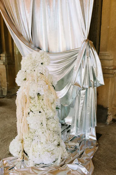 Wedding ceremony backdrop with a silver draped backdrop and white florals, pampas grass accents, framed by stone columns outdoors