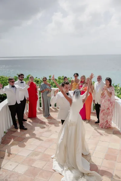 Just married celebration with wedding guests cheering, the white-tuxedo groom and bride gathered on an oceanfront terrace against sky and greenery.