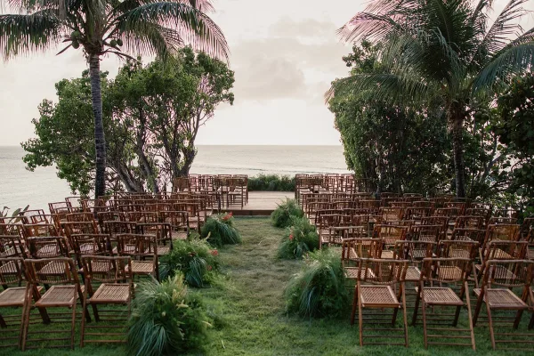 Ceremony setup highlights elegant ceremony aisle design with bamboo chairs lining a natural grass aisle, set before an ocean view and palm trees.