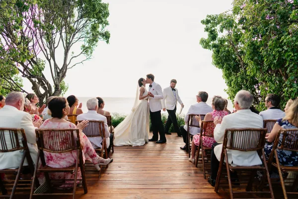 Ceremony kiss with wedding guests applauding around them, her veil catching light on a wooden deck overlooking the ocean and trees.