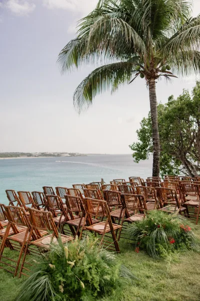 Beach ceremony setup with bamboo wedding chairs facing the ocean, flanked by aisle greenery on a sunlit coastal lawn.