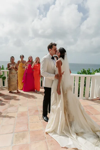 Wedding kiss portrait as the couple shares a kiss with wedding party cheering, champagne glasses raised, on a terrace by the ocean and white railing.