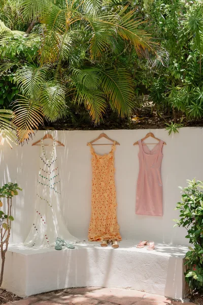 Bridesmaid dresses, a row of mismatched bridesmaid dresses on hangers, drape against a white stucco wall edged with palm leaves and greenery.