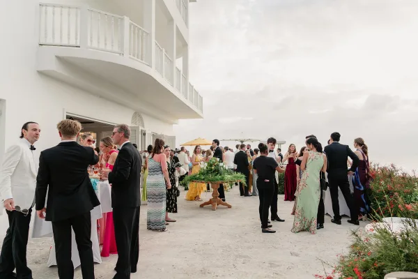 Wedding cocktail hour with guests mingling at an outdoor cocktail reception on a terrace, cocktail tables dot an ocean-view patio by a white facade.