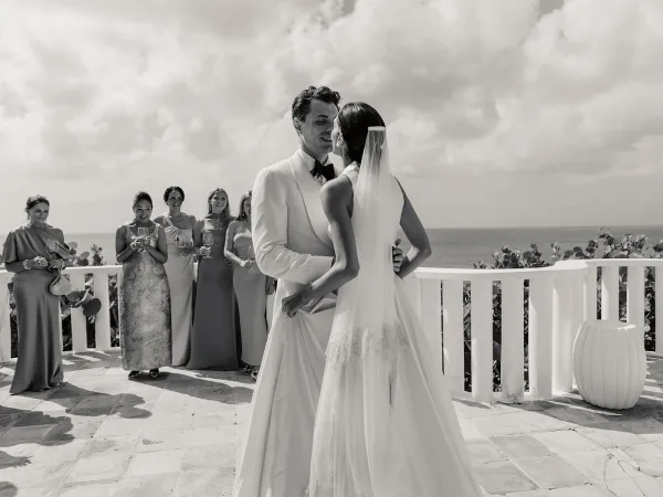 Couple portrait of a bride and groom embrace on a terrace overlooking the ocean, her bridal veil catching light by the white railing.