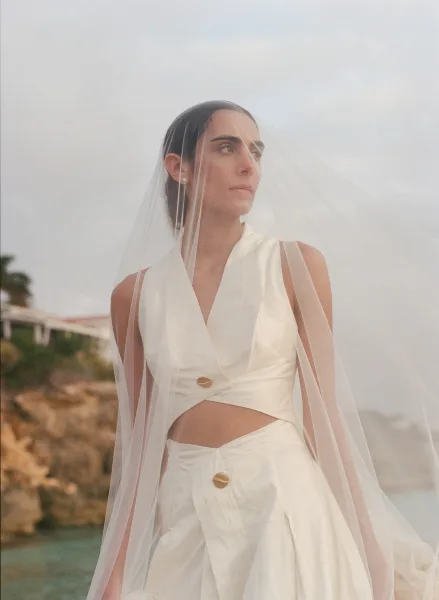 Bridal portrait of a bride under veil in a two-piece dress, framed by the ocean and rocky cliffs beneath a cloudy coastal sky.