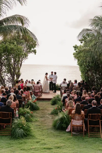 Ceremony moment during an outdoor wedding ceremony as the bride in a flowing veil and groom stand on an oceanfront lawn framed by palm trees.
