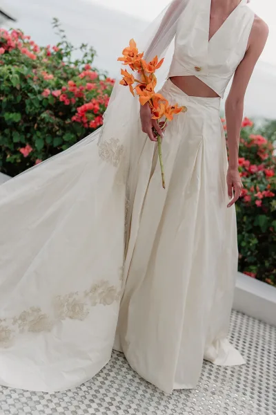 Wedding dress close-up of a two-piece wedding dress as the bride holds an orange orchid bouquet against a garden and ocean backdrop.