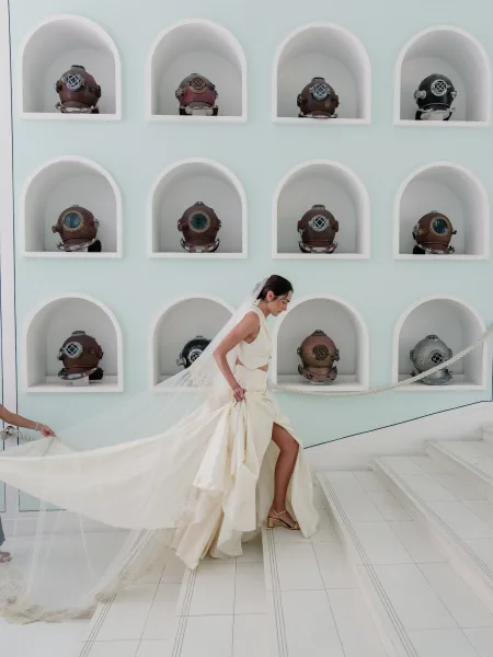 Bridal portrait of a bride walking stairs, her long veil trailing over white tile stairs, past arched niches displaying vintage diving helmets.