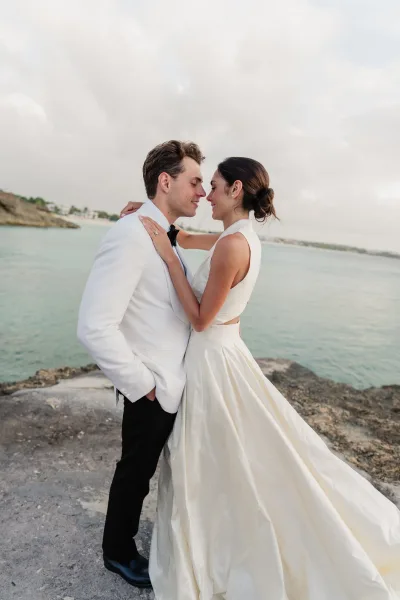 Couple portrait at a rocky ocean shore under a cloudy sky, a waterfront wedding portrait with tux-clad groom in a bow tie and a bride in a satin gown