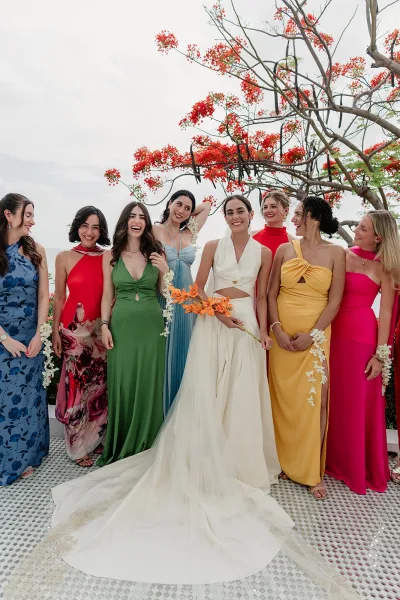 Bride and bridesmaids in colorful bridesmaid dresses laugh together, wearing orchid leis beside a flowering tree under a cloudy sky.