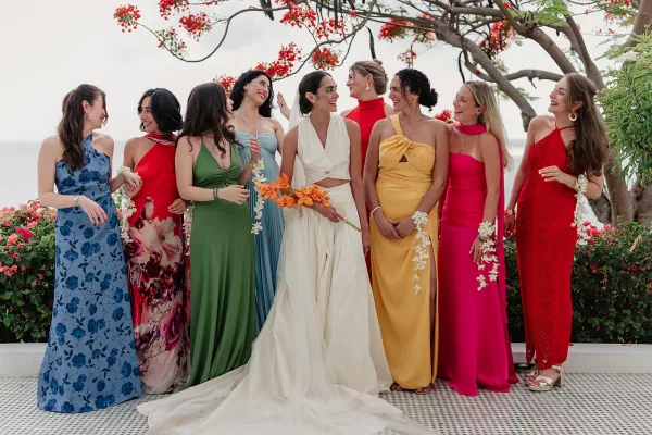 Bride with bridesmaids in colorful bridesmaid dresses pose by an ocean view, the bride holding an orange orchid bouquet.