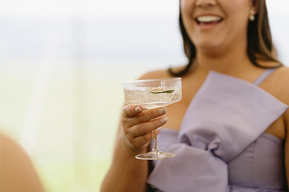 Bridesmaid toast as she lifts a cocktail coupe glass with lime garnish, wearing a lavender bow dress and pearl studs in outdoor light