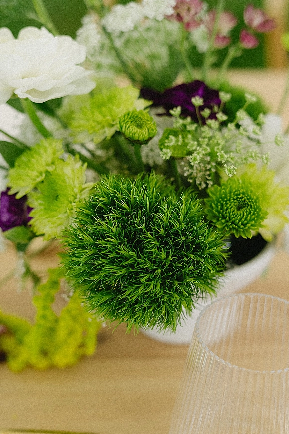 Wedding centerpiece with a green wedding centerpiece of white blooms, lime pom pom flowers, purple accents, and a ribbed glass votive on wood table