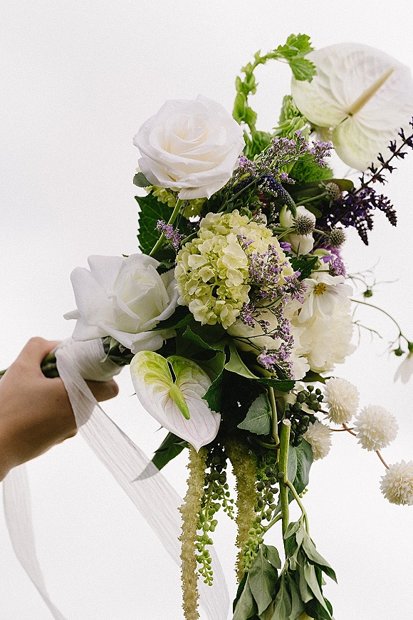 Bridal bouquet with white roses and anthurium, hydrangea, thistle, and hanging greenery, wrapped in ribbon and held against a white backdrop