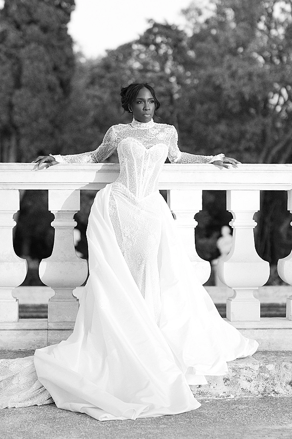 Bridal portrait in a high neck wedding dress with long sleeve lace, beaded bodice, and tulle overskirt on a stone terrace balustrade