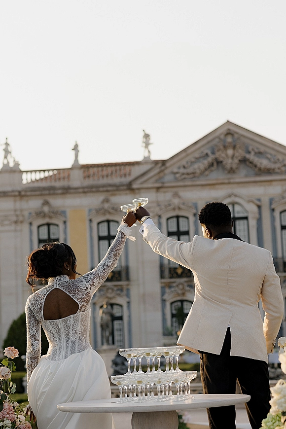 Wedding champagne toast as bride in long sleeve lace dress and groom in white dinner jacket clink coupes beside a tower at an estate facade outdoors