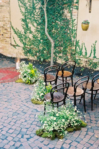 Ceremony aisle design with outdoor ceremony seating framed by wildflower clusters in a cobblestone courtyard against an ivy-covered brick wall.