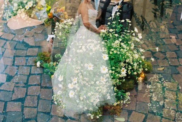 Ceremony moment with bride and groom holding hands, framed by daisy florals on cobblestone amid garden greenery.