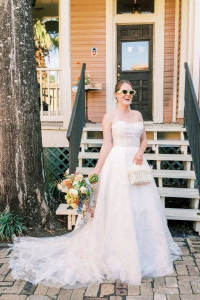 Bridal portrait of a bride on stairs, laughing in cat-eye sunglasses on wooden porch steps beside a black door and wooden house facade.