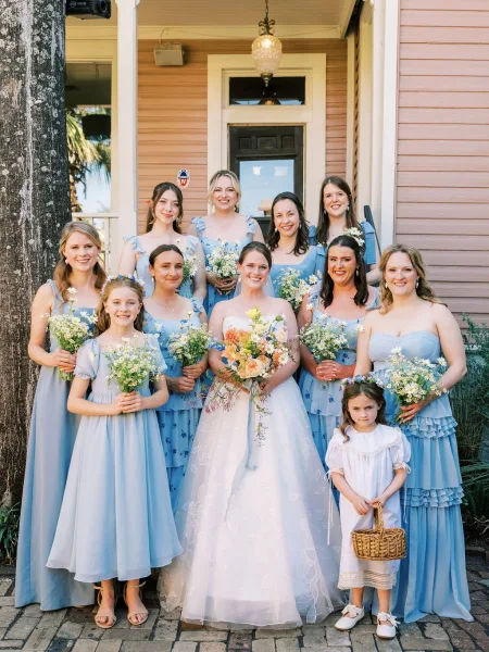 Bridal party portrait featuring the bride with bridesmaids and flower girls on a brick walkway by a house porch, in light blue bridesmaid dresses.