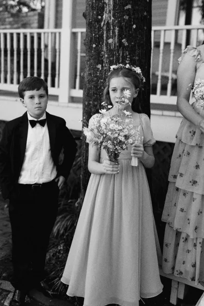 Flower girl portrait with ring bearer in tux beside her, posed by a porch railing and house exterior with a tree trunk and leafy plants.