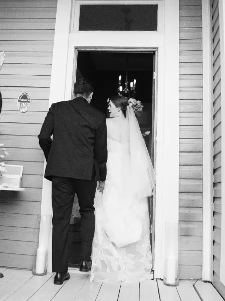 Couple entrance through a rustic wood doorway, the bride entering venue with a flowing tulle veil as they step from porch to a dim interior.
