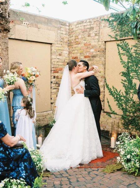 Wedding first kiss as the bride and groom embrace beneath ivy on a brick courtyard wall, bridesmaid bouquets framing the moment.