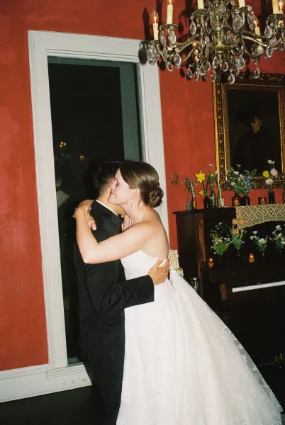 First dance as the bride and groom embrace beneath a crystal chandelier, in a room with a red wall and a framed painting.